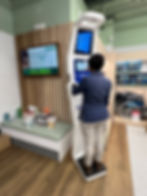 Person using an Abby Health Kiosk in a store, surrounded by wellness products. Digital display shows health information. Warm, inviting setting.