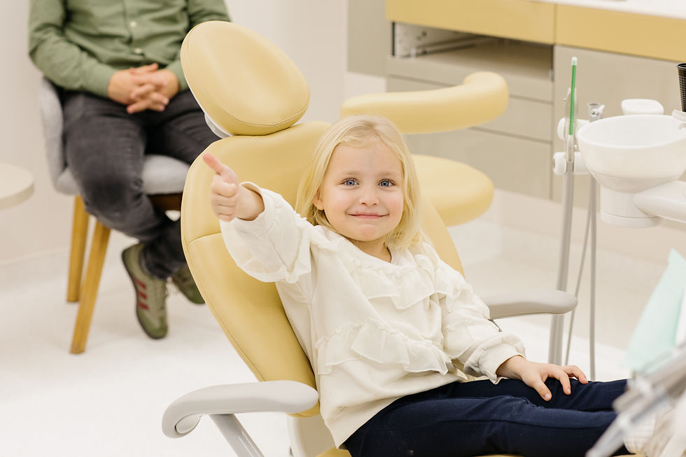 Fetiță blondă, zâmbitoare, într-un scaun galben de dentist, oferă un semn de "ok" cu degetul mare ridicat. Fundal alb, bărbat în verde. Emoție de încredere.