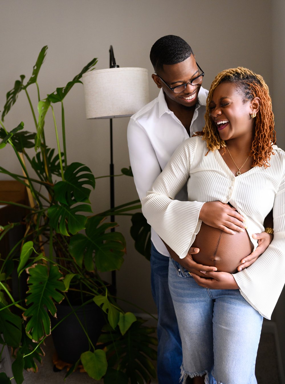 Expecting couple laughing together, hands on her bare belly, beside a monstera plant