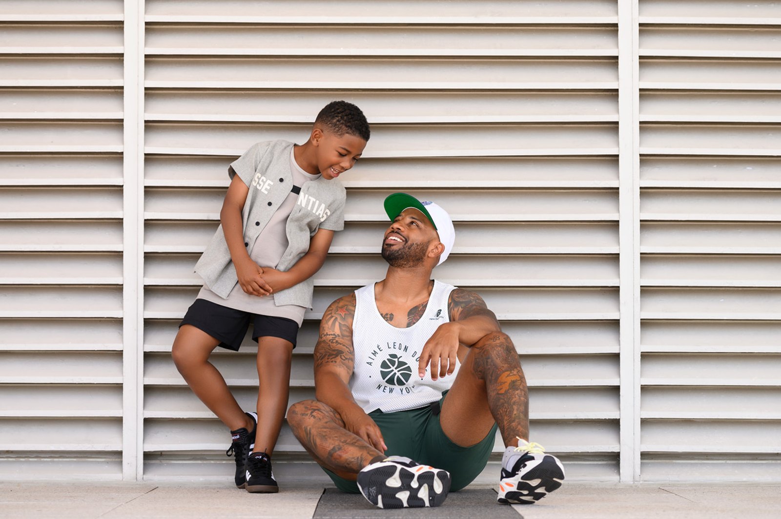 A smiling boy leaning on a louvered wall, looking down at his laughing father seated beside him