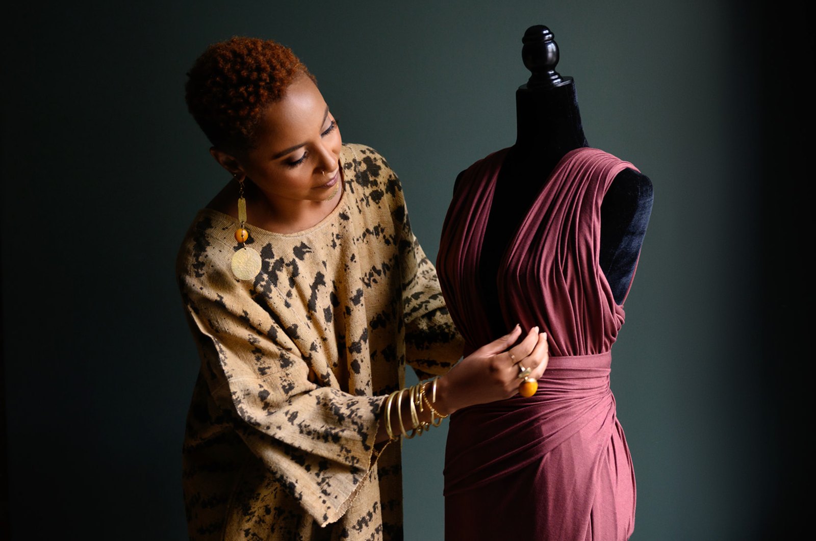 A designer in a printed tunic adjusting a draped plum dress on a dress form against a dark green wall