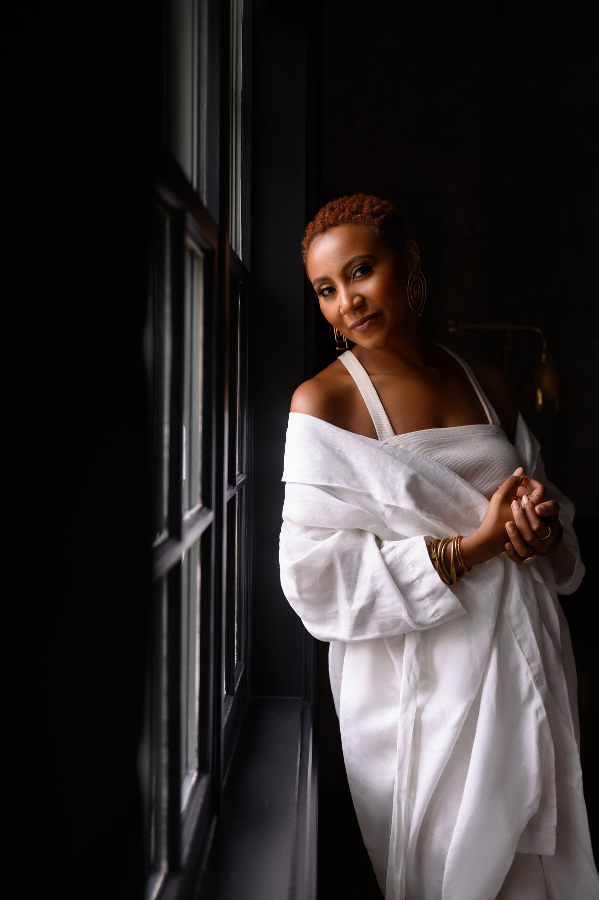 Woman in a white linen wrap leaning against a dark window frame, softly smiling