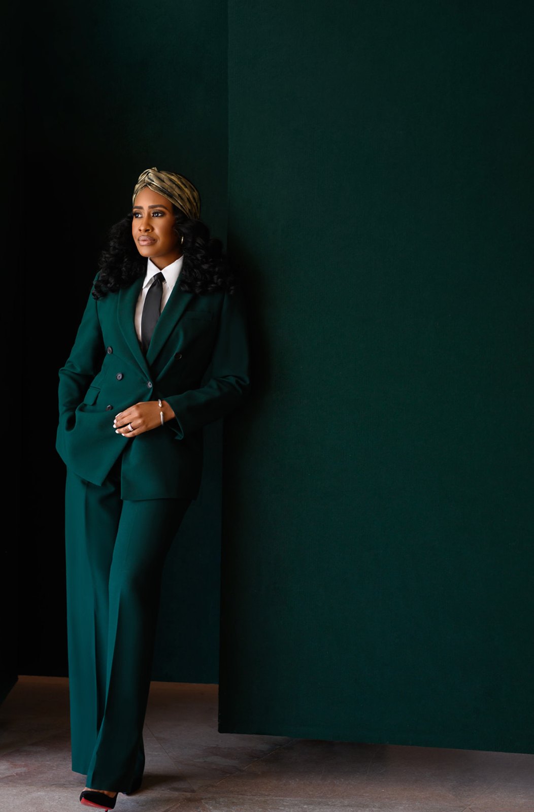 Woman in a tailored emerald double-breasted suit and gold headwrap leaning against a deep green wall