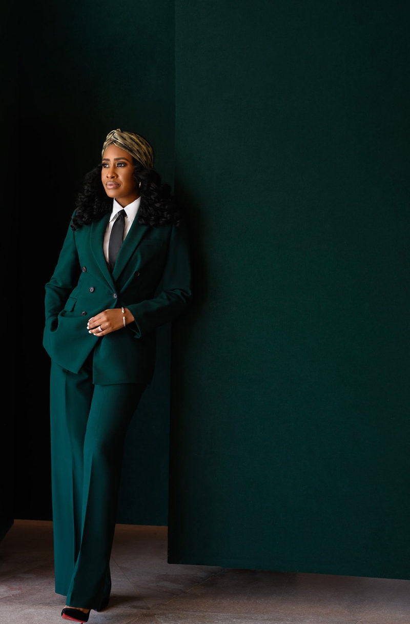 Woman in a tailored emerald double-breasted suit and gold headwrap leaning against a deep green wall