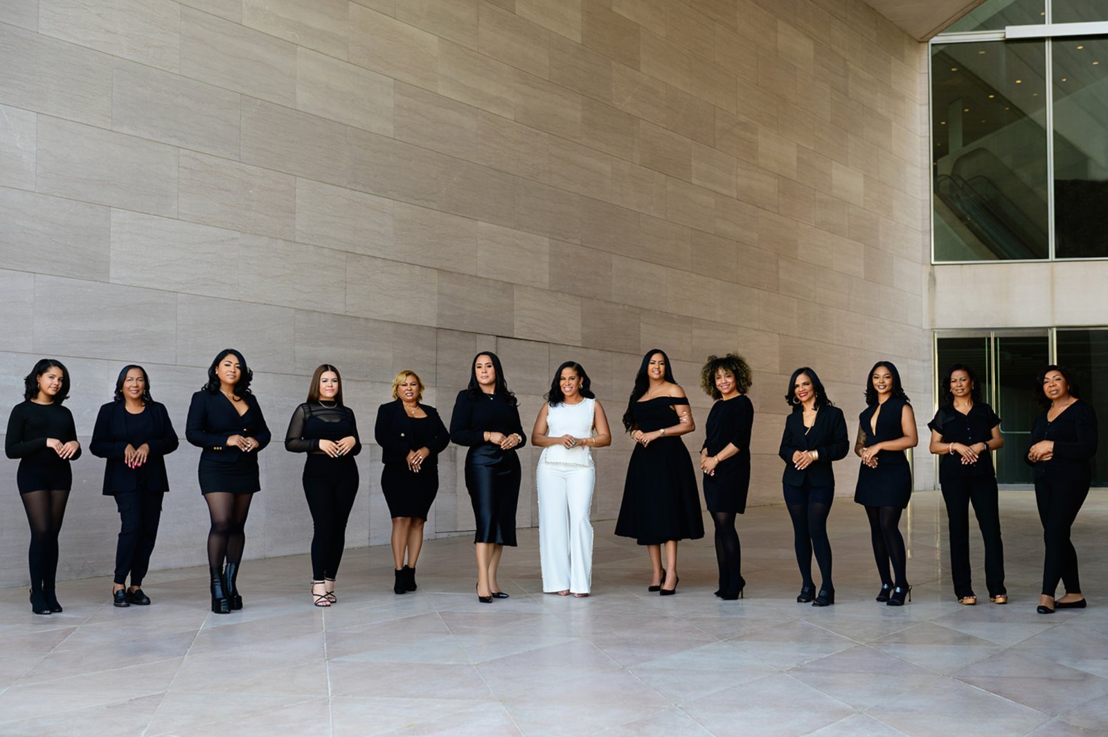 Thirteen women in coordinated black outfits, with their leader in white at center, standing in a line before a stone wall