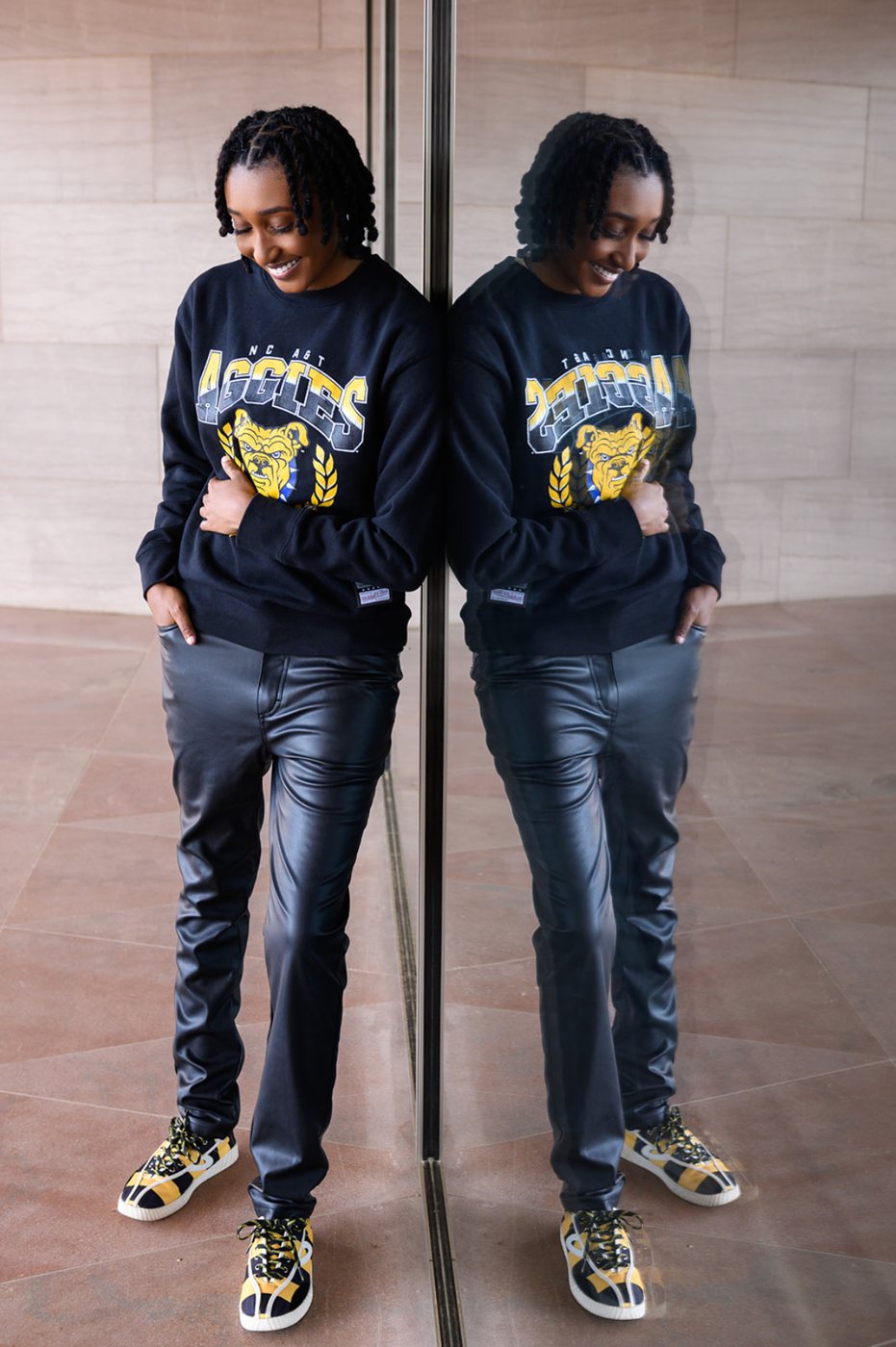 Senior in an Aggies sweatshirt and leather pants leaning on a glass wall beside her reflection