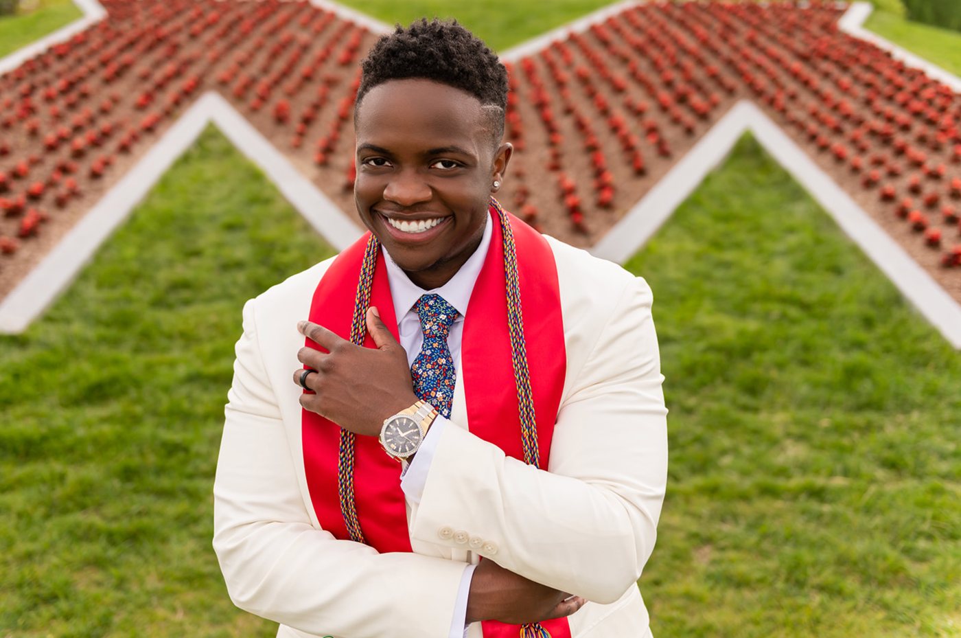 Graduate in an ivory suit and red stole with cords, smiling in front of a red flower garden