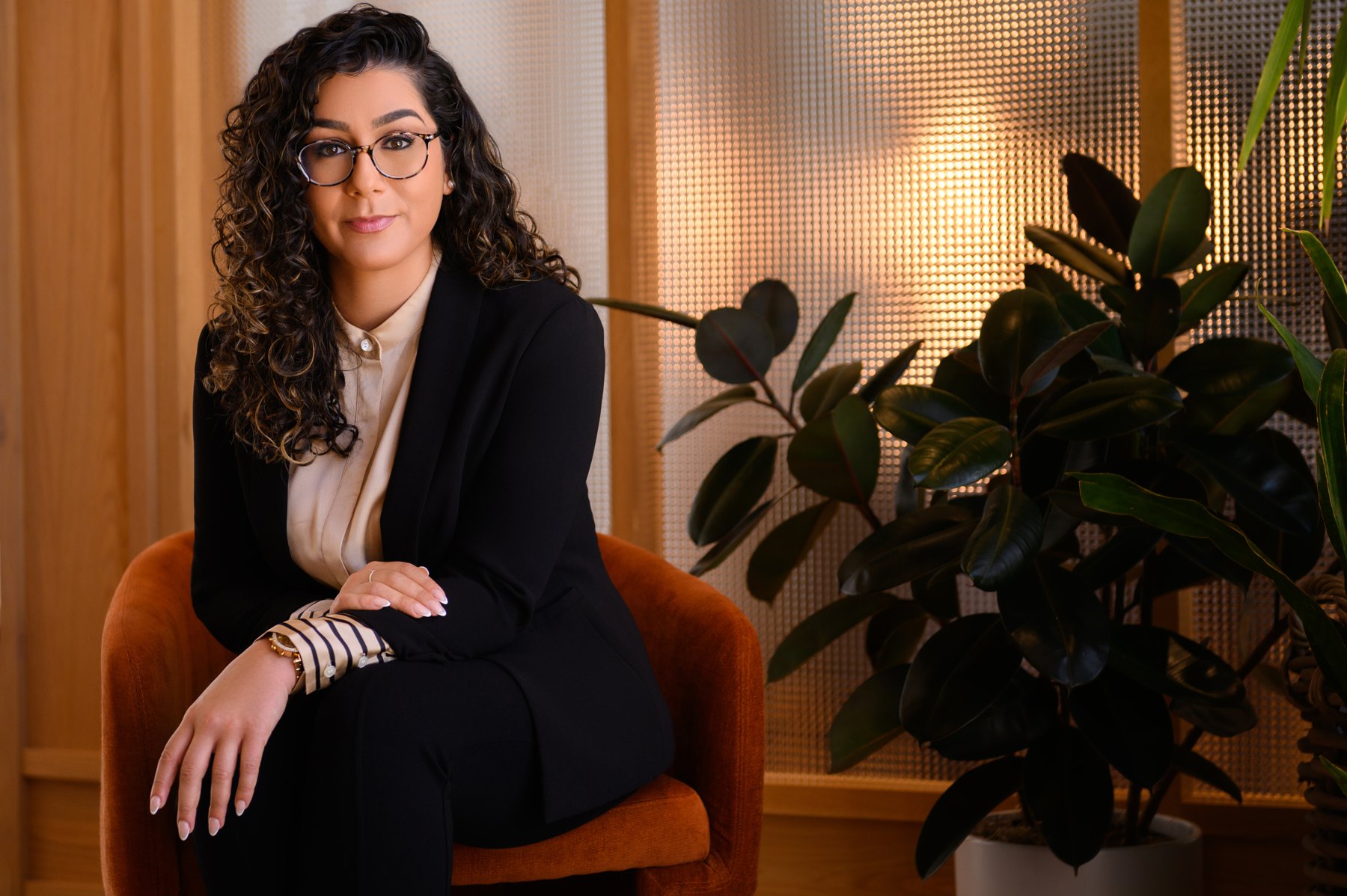 Professional in glasses and a black blazer seated on a rust velvet chair beside a large plant
