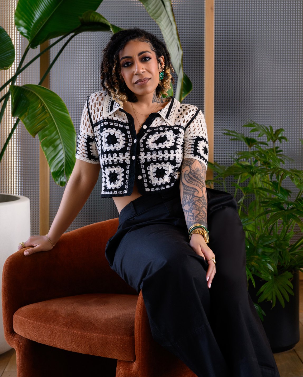Ashleigh Bing seated on a rust velvet chair in a crochet top, surrounded by green plants