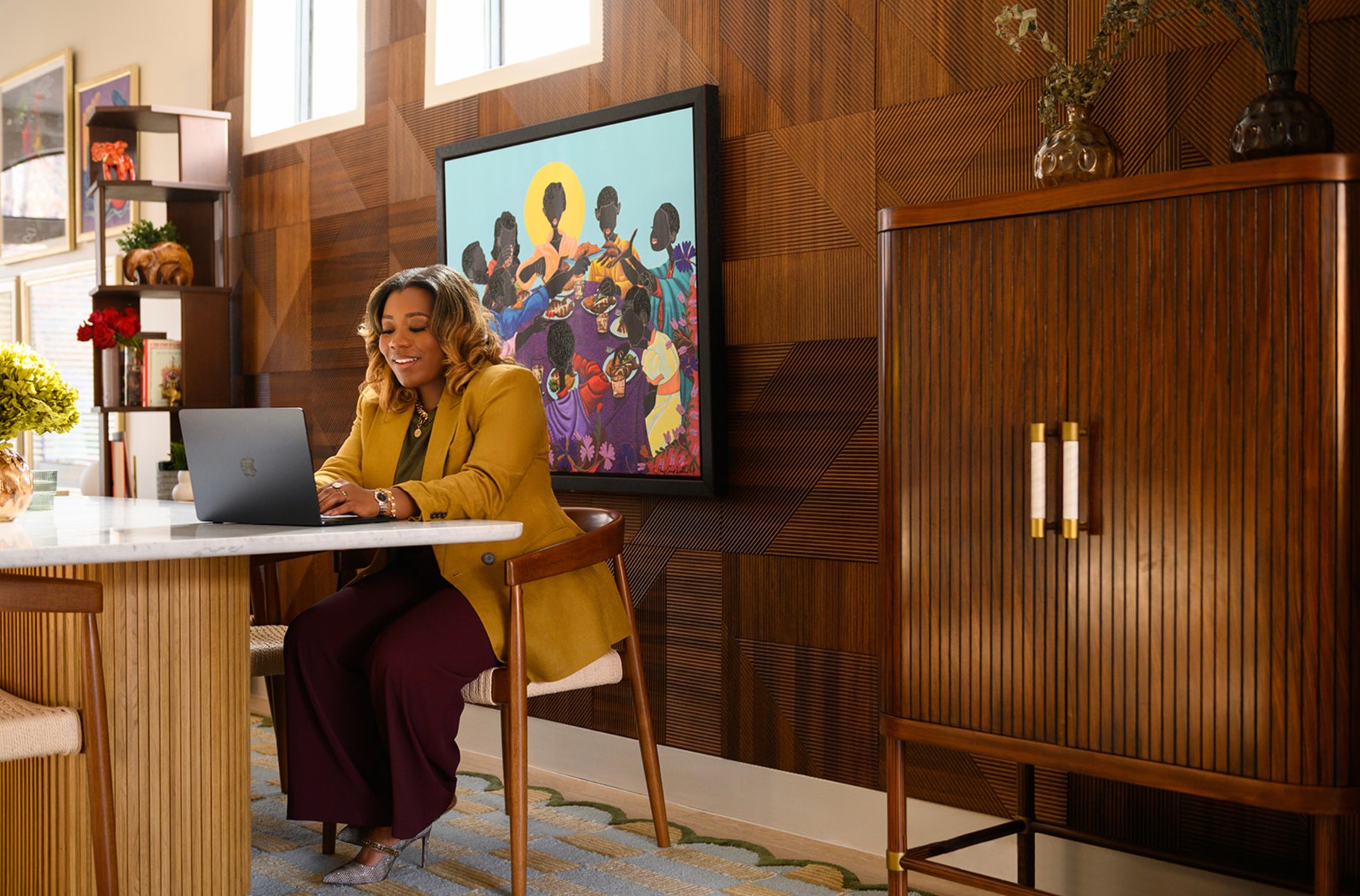 Business owner in a mustard blazer smiling at her laptop at a marble table in a warm wood-paneled office with colorful art