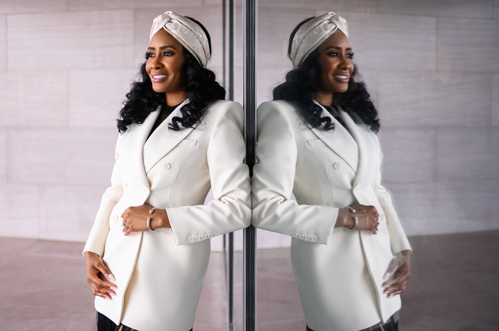Woman in an ivory blazer and headwrap leaning on a glass wall, her reflection beside her