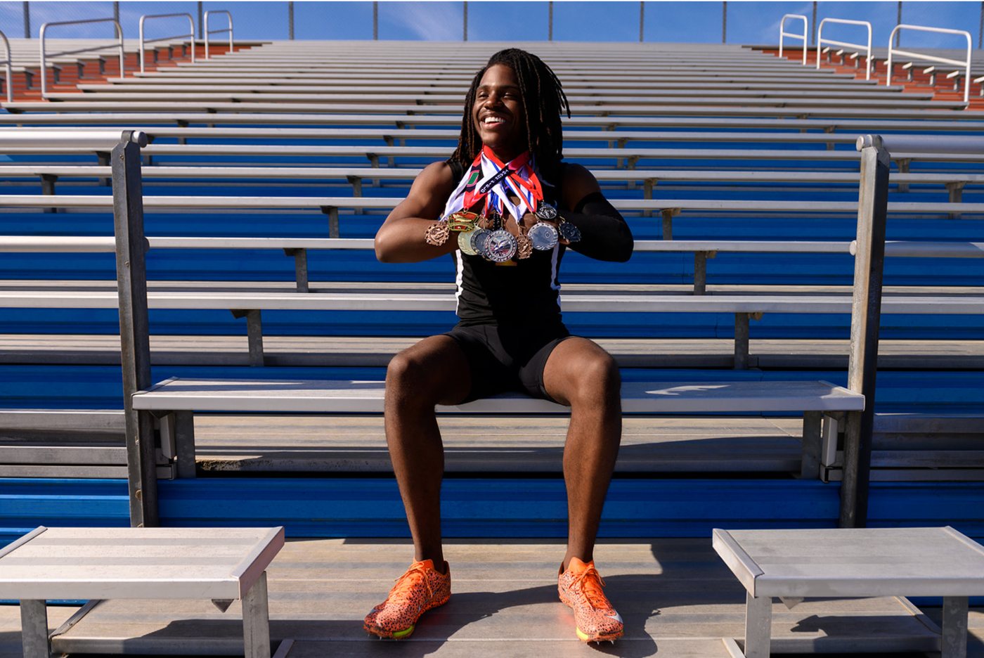 Track athlete grinning on blue bleachers, holding up a chest full of medals