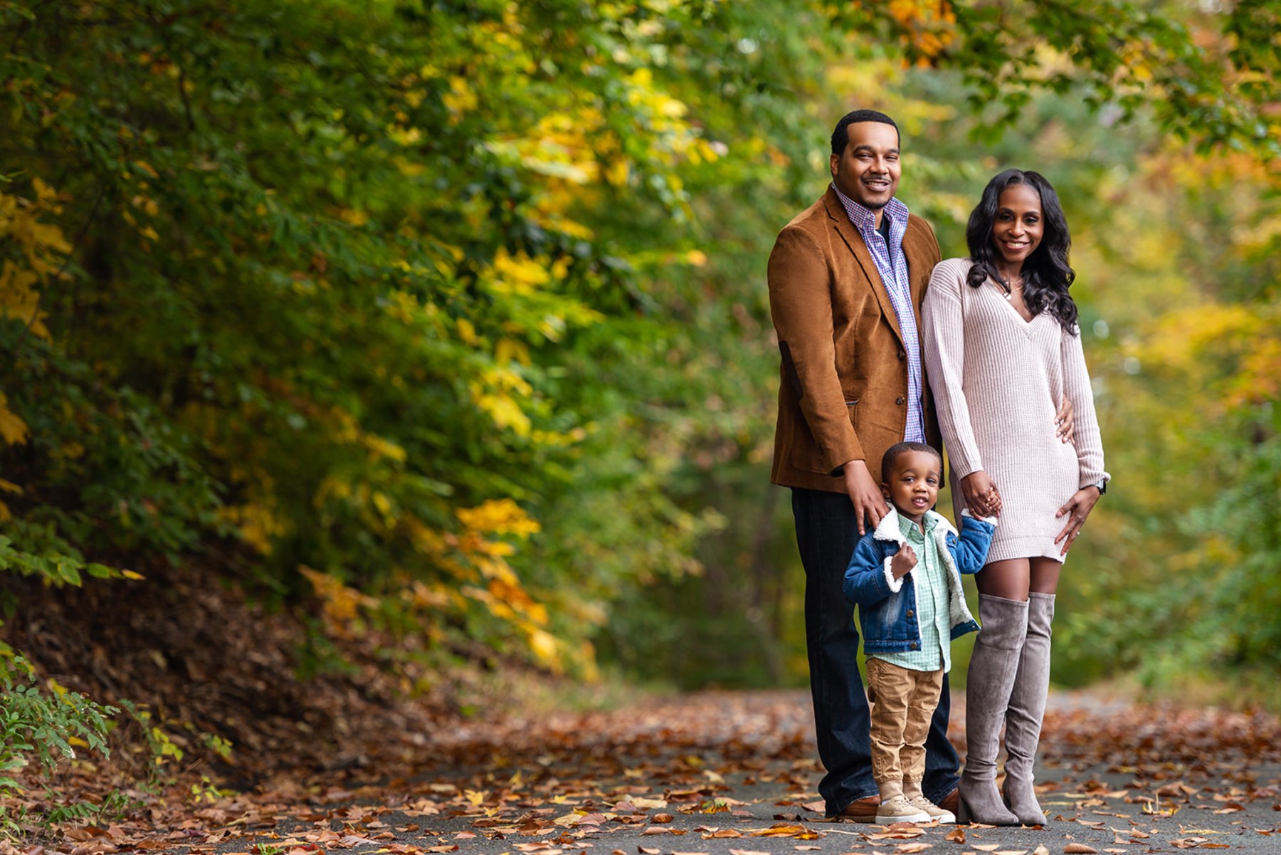 Parents and their toddler son smiling on a leaf-covered path beneath bright autumn trees