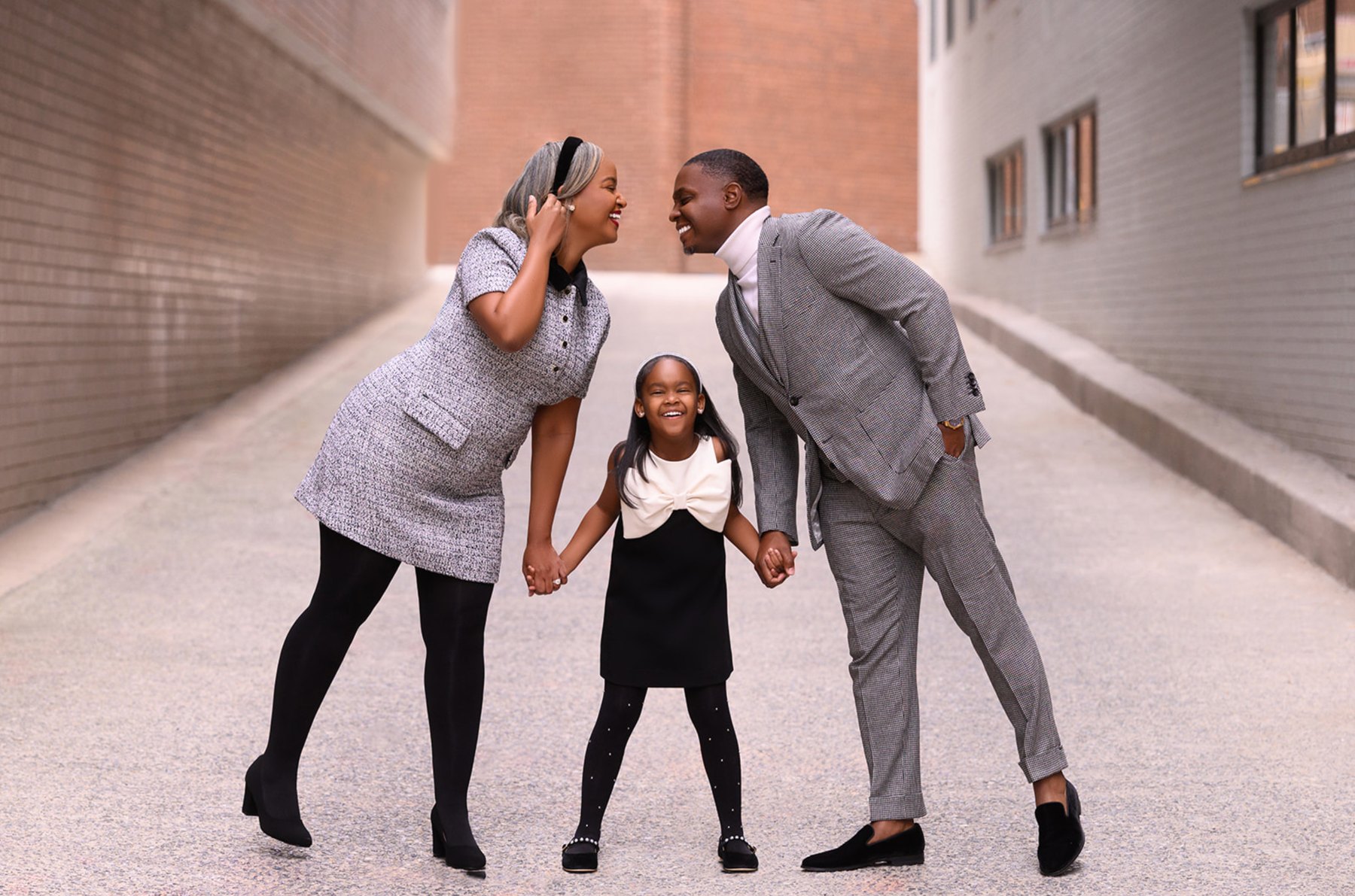 Parents leaning in nose to nose, laughing, while holding hands with their smiling daughter in a city alley