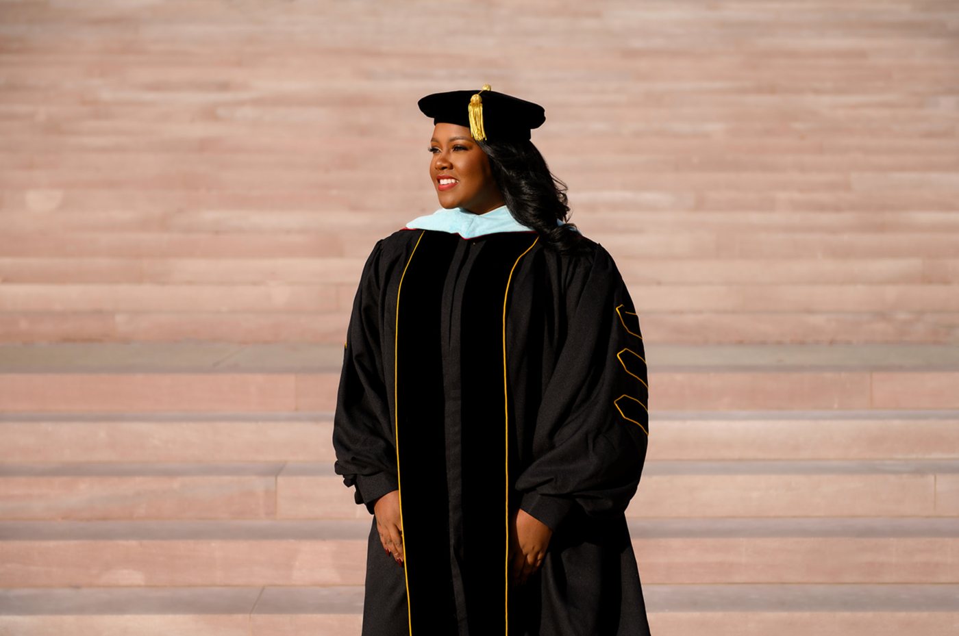 Doctoral graduate in velvet-trimmed robes and tam standing on sunlit stone steps