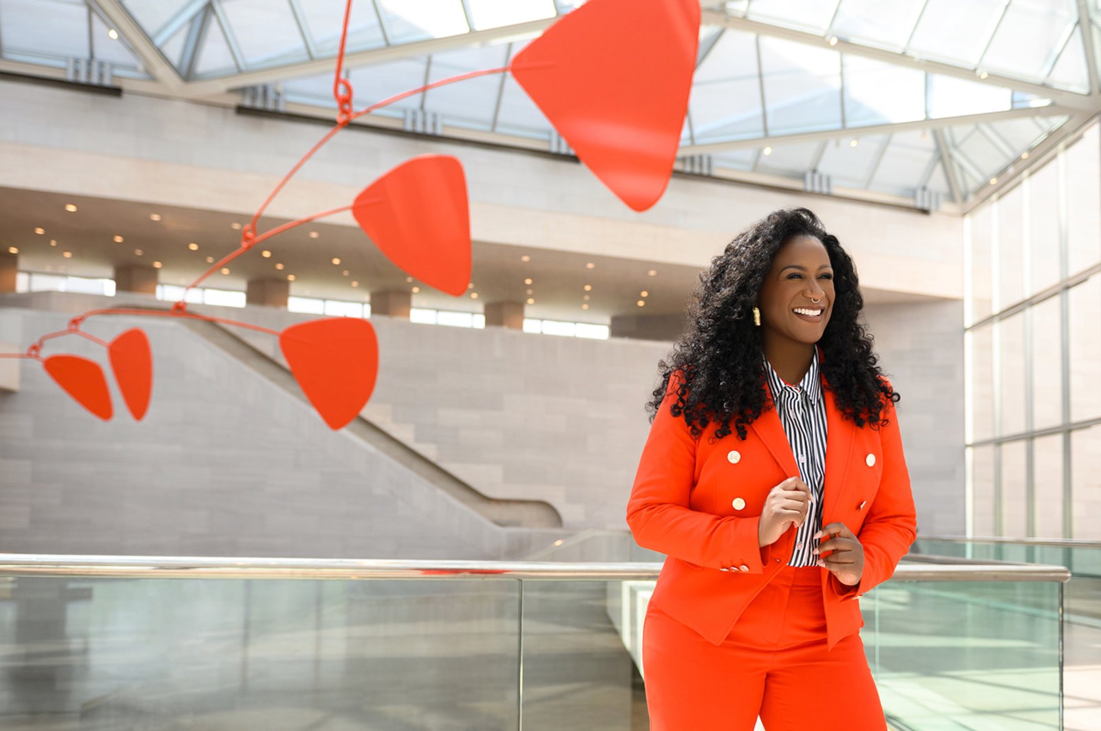 Smiling woman in a bright orange suit beneath a red hanging mobile in a museum atrium