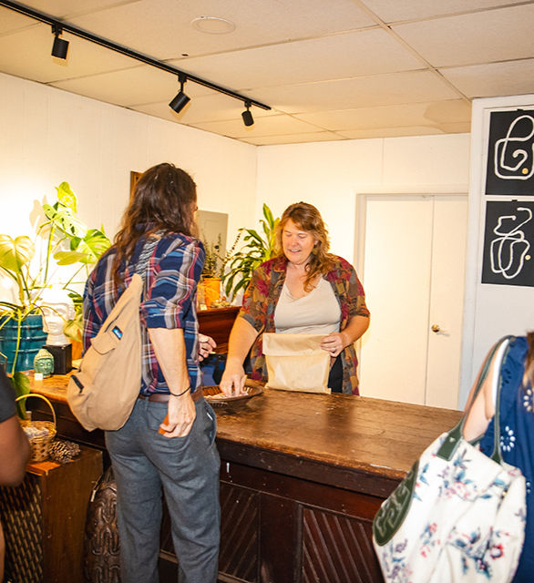 A woman hands a paper bag to a man across a wooden counter in a plant-decorated shop. Both are smiling, creating a friendly atmosphere.