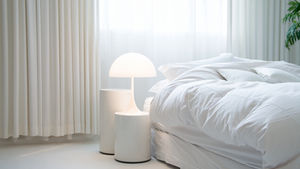 Fluffy, clean, white bed surrounded by white curtains and a umbrella bedside lamp on a table