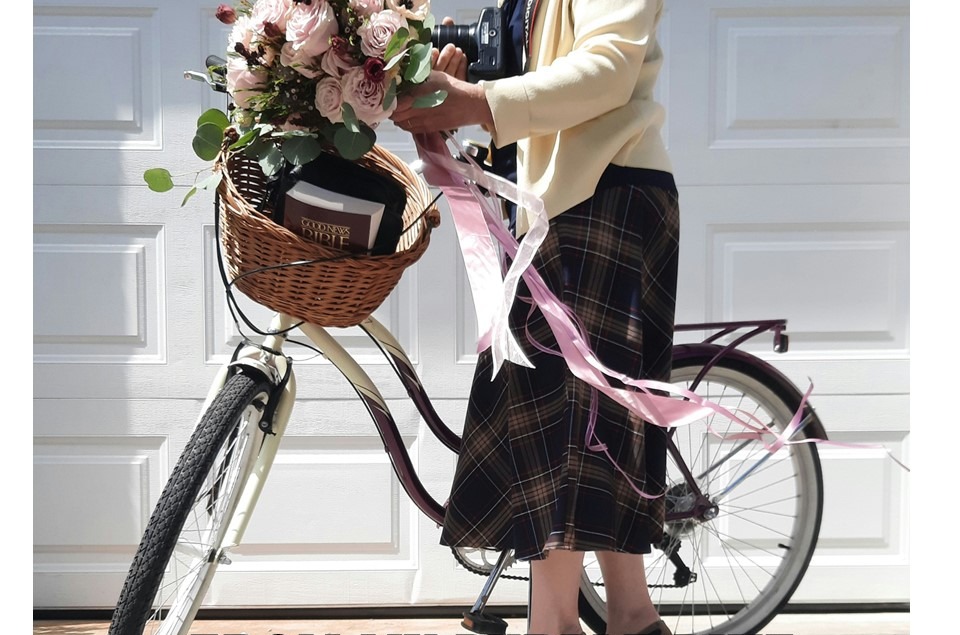 Beata na fioletowym rowerze – w koszyku Biblia, aparat fotograficzny i przepiękny bukiet kwiatów z długimi, falującymi wstążkami. Symbol mojej drogi: Słowo Boże, uwiecznianie Bożych darów i wdzięczność za piękno stworzenia.