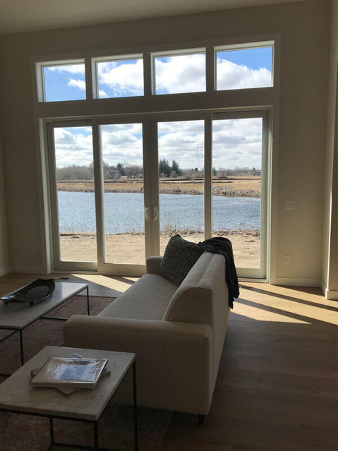 Light tan colored living room with a cozy, tan couch. Faces a large, glass set of doors and windows looking out at a lake. Sunshine peaks through the windows.