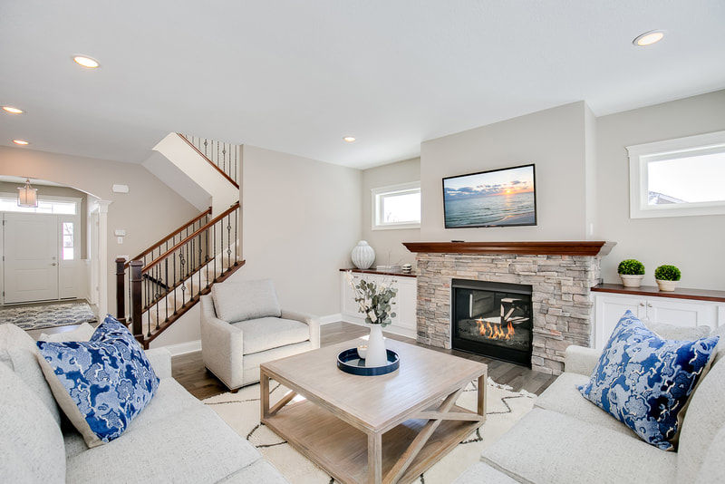 View of living room and foyer of the two story home in Rockford, MN. Includes a stone fireplace with a TV mounted above it, white upholstered furniture and square coffee table in the center. Photo shows open staircase with wrought iron spindles, oak handrail , and oak newel posts, leading to the second floor.