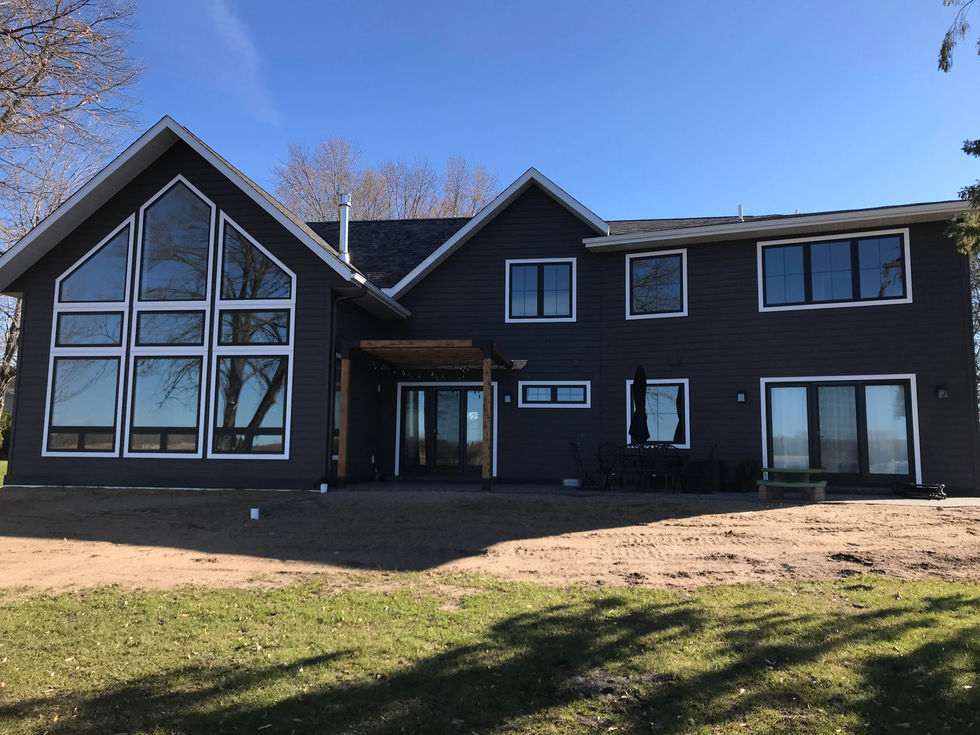Lakeside view of home. Former three-season porch was replaced with a new expansive great room featuring twelve-foot side walls, twenty-foot high vaulted ceiling and tall-wall with peak pentagon window that is flanked by trapezoid windows. Two-story addition on existing rambler includes numerous large windows overlooking lake. Dark brown siding, white windows.