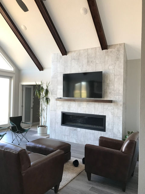 Image of a living room with a large statement fireplace. The fireplace is white with a TV mounted near the top. The living room has light gray walls with dark brown leather couches and dark brown wooden beams running across the ceiling.