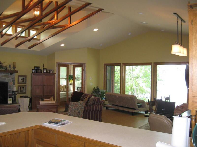 Image of living room after remodel. Dormer with three windows were added, exposing existing roof trusses wrapped in copper. Fireplace wall was removed to increase size of living room and to make room for more windows. New, larger windows replace old windows; pendant lighting added over dining table. Fresh light green paint finishes it off.
