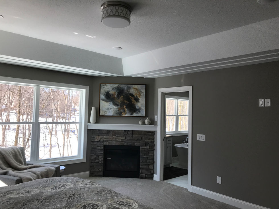 Bedroom suite with tray ceiling, dark gray walls, white baseboard and cove moulding, corner gas fireplace with stone surround and white mantel, and a doorway to the primary bathroom.