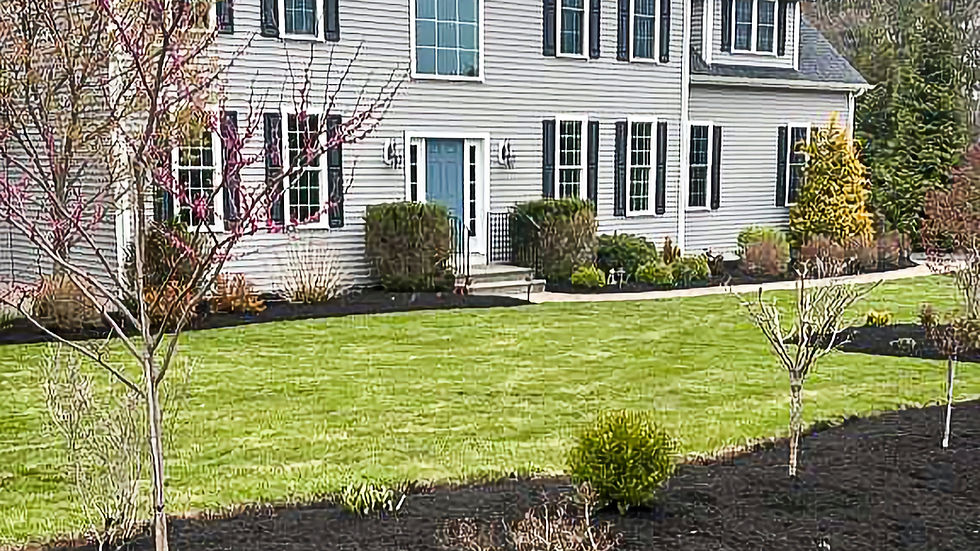 Large gray house with black shutters, manicured lawn, fresh mulch, and young trees. Overcast sky and lush greenery in the background.