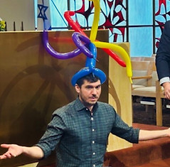Rabbi Daniel Reichenbach wears a colorful balloon crown while another man laughs during a playful synagogue program.
