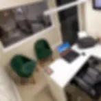 Escritório com mesa branca, notebook, impressora e poltronas verdes.