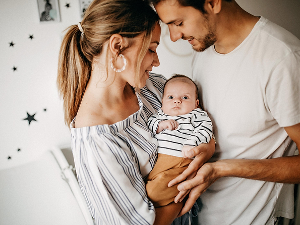 alt="mother holding baby in her arms and father on looking, man in white t-shirt, baby wearing black and white striped shirt and brown pants, white wall with black stars in the background"
