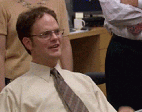 Man in glasses and a tie smiles and leans back in an office setting, surrounded by people. The mood is lighthearted and relaxed.