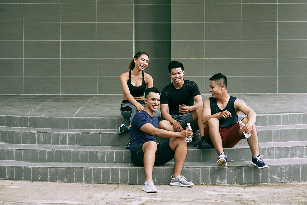 Four healthy male and female sit on gray steps, laughing and chatting. They're in casual workout gear with water bottles, set against a tiled wall.