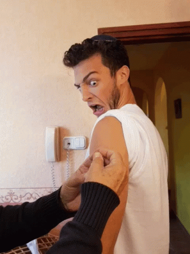 Man with surprised expression receiving a shot in his arm indoors. He wears a white shirt; the background features a wall phone and arched doorway.