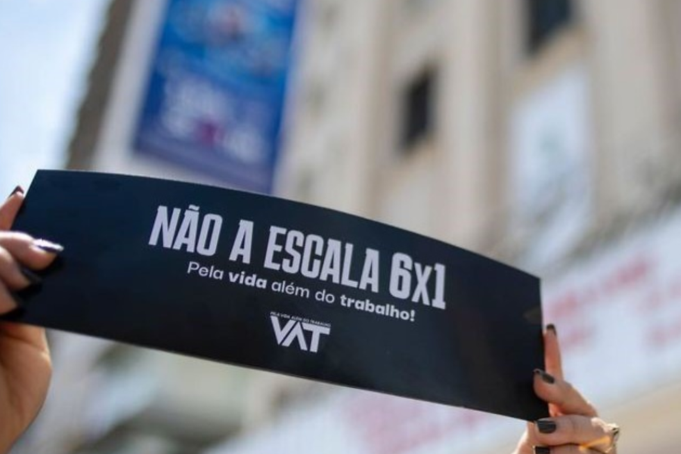 Mãos seguram placa preta com texto "NÃO A ESCALA 6x1. Pela vida além do trabalho". Fundo desfocado com prédio. Tom de protesto.