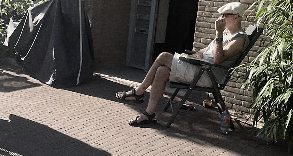 Hombre con gorra y anteojos de sol, sentado en una silla plegable en la calle, mirando a dos personas que pasan, con una pared de ladrillos y una planta de fondo, en una vereda empedrada de Amsterdam.