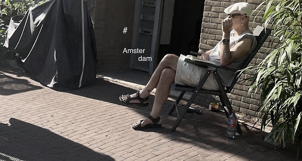 Hombre con gorra y anteojos de sol, sentado en una silla plegable en la calle, mirando a dos personas que pasan, con una pared de ladrillos y una planta de fondo, en una vereda empedrada de Amsterdam.