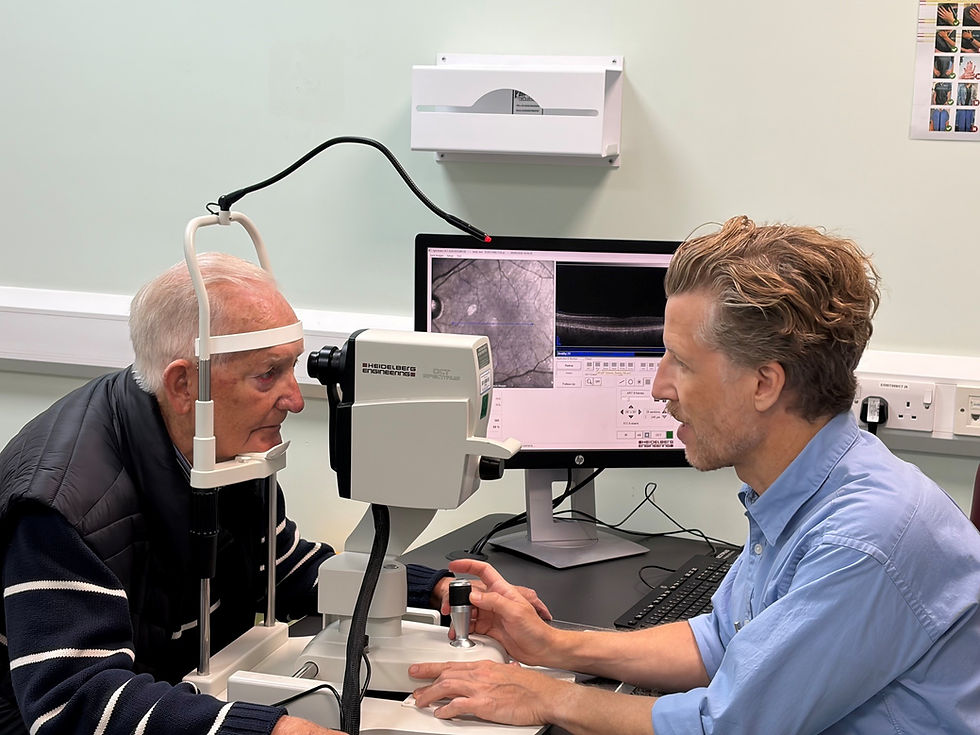 Colour photo of a patient with his face leaning into an OCT retinal imaging device. Opposite sits an ophthalmologist with computer screen next to him showing a black and white retinal image