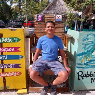 A teenage boy sitting on an oversized wooden chair next to a colorful mile marker destination sign at Robbie's Marina.