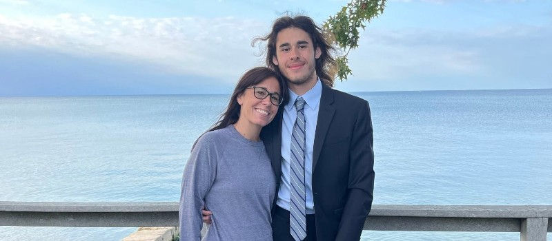 Melissa hugs Jacob in a suit by the calm ocean, with blue sky and a wooden railing behind them.