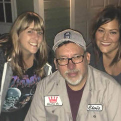 A close-up selfie of Mara and two friends smiling together indoors. On the left is a woman with blonde hair wearing a dark graphic t-shirt. In the center is a man with a beard, glasses, and a grey baseball cap, wearing a light tan button-up work shirt with patches. To the right is Mara