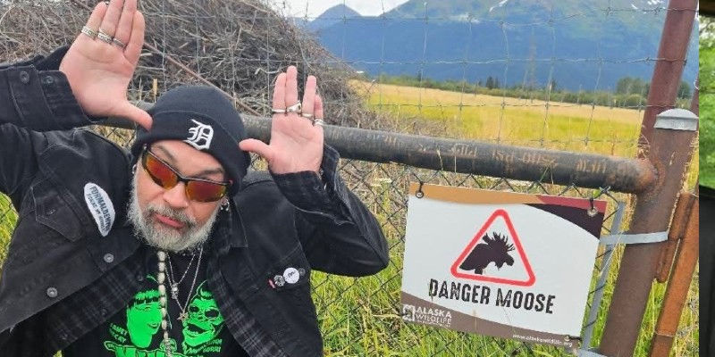 J Michael, in sunglasses poses like antlers beside a DANGER MOOSE sign in a grassy Alaska field.