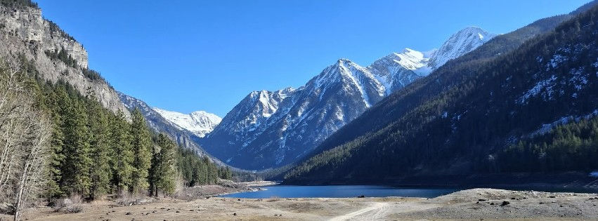 Snow-capped mountains rise above a blue lake and evergreen forest under a clear sky in a calm alpine valley.