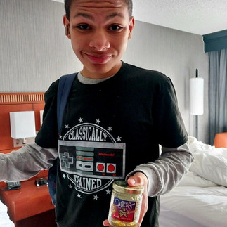A close-up of a teenage boy smiling in a hotel room, wearing a black graphic t-shirt that reads "Classically Trained" with a retro Nintendo controller design. He is holding up a small glass jar of food toward the camera.
