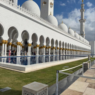 A perspective shot looking down the side of the grand white marble arcade at the Sheikh Zayed Grand Mosque, with its columns and white domes perfectly reflected in a long, calm blue pool in the foreground under a cloudy blue sky.