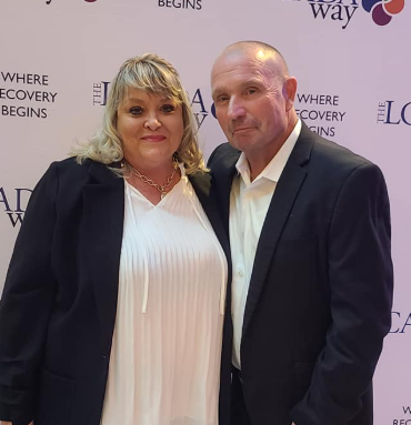 A formal portrait of Michele Taft and her husband Martin standing together at an event. Michele is smiling, wearing a white pleated top and a black blazer. Martin is beside her in a dark suit jacket and white button-down shirt. The background features a light pink backdrop with the words "Where Recovery Begins.