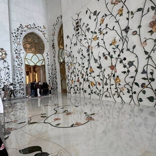 A detailed interior view of the Sheikh Zayed Grand Mosque, showcasing the smooth, polished white marble floor and walls adorned with intricate, winding floral and vine mosaic inlays made of colorful semi-precious stones.