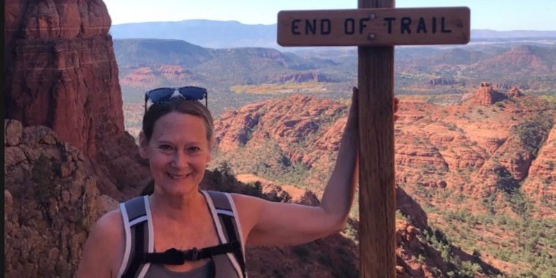 Smiling, Shari beside an END OF TRAIL sign, posing on a rocky canyon overlook with red cliffs and mountains.